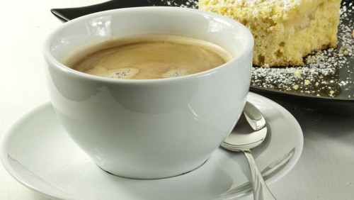 Das Bild einer weißen Tasse mit Kaffee und einem kleinen Löffel. Im Hintergrund ist ein angebissenes Stück Kuchen mit Puderzucker zu sehen.