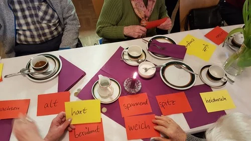 Ein Foto von einem gedeckten Tisch mit Kaffeegeschirr und bunten Papierkarten, auf denen Worte stehen. Am Tisch sitzen mehrere Senior:innen. Das Foto ist von oben aufgenommen.