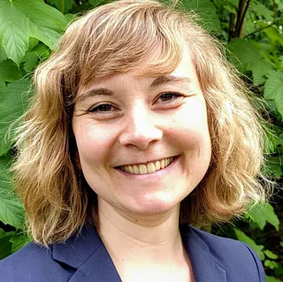 Ein Foto von Ellen Schlager vor grünem Hintergrund. Sie trägt einen blauen Blazer und lächelt direkt in die Kamera.