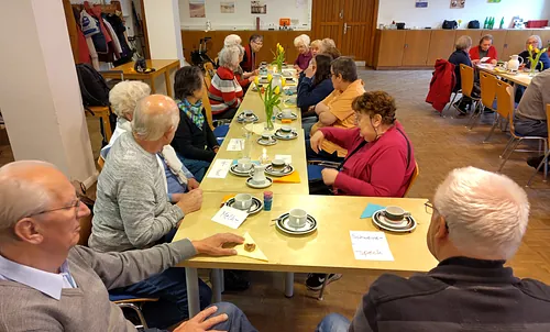 Ein Foto von einem gedeckten Tisch mit Kaffeegeschirr und bunten Papierkarten, auf denen Worte stehen. Am Tisch sitzen mehrere Senior:innen. Das Foto ist von oben aufgenommen.
