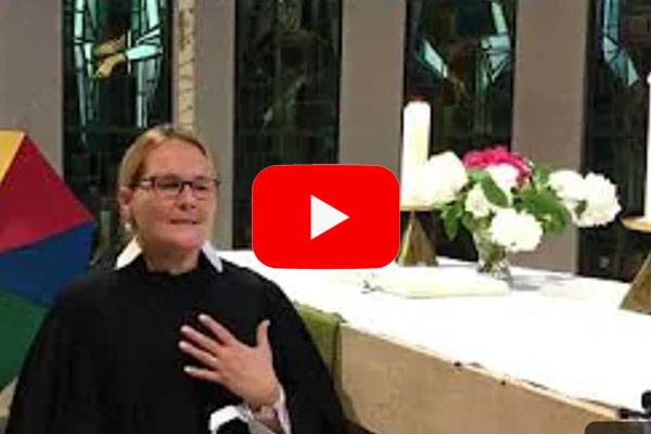 Startbild des Youtubevideos: Pastorin Saskia Tluczykonz im Talar vor dem Altar, der mit Blumen und Kerzen geschmückt ist.